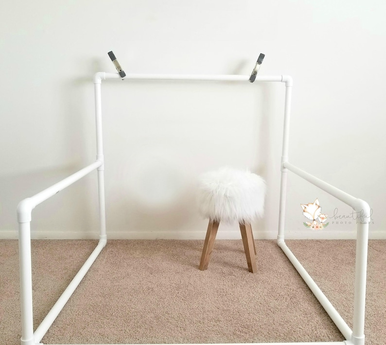 May include: White PVC pipe backdrop stand with a white faux fur stool in the center. The stand is assembled in a rectangular shape with two horizontal bars and two vertical bars.