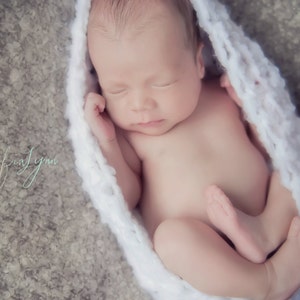 May include: A newborn baby is sleeping soundly in a white crocheted wrap. The baby is lying on its back with its arms tucked in and its legs curled up. The baby's face is peaceful and serene.