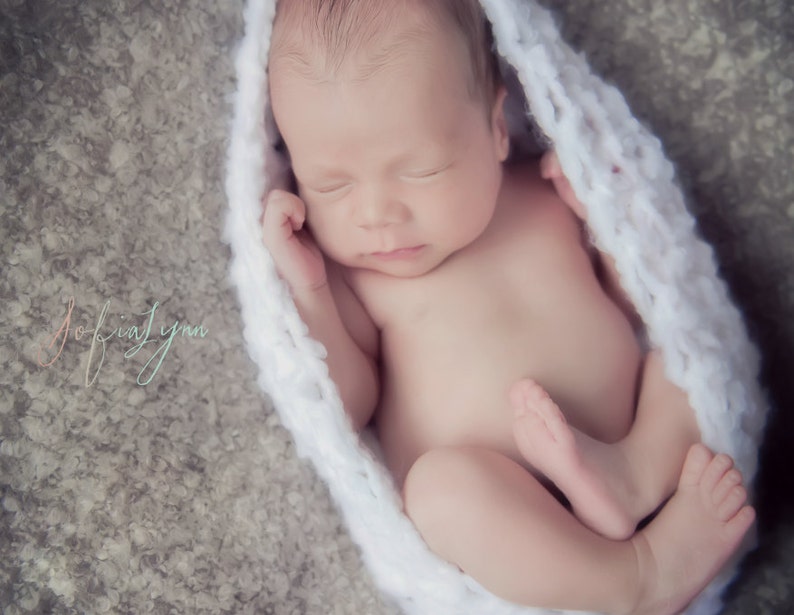 May include: A newborn baby is sleeping soundly in a white crocheted wrap. The baby is lying on its back with its arms tucked in and its legs curled up. The baby's face is peaceful and serene.