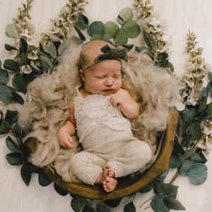 May include: A newborn baby wearing a white crocheted outfit and a green headband, sleeping in a wooden bowl surrounded by green leaves and white flowers.