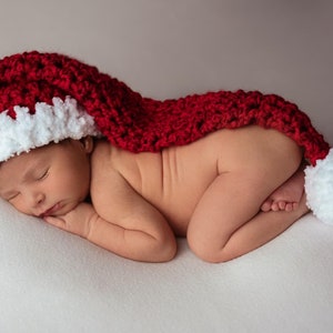 May include: A newborn baby wearing a red and white crocheted Santa hat, sleeping on a white blanket.