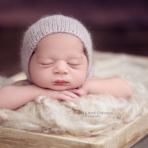 Puede incluir: Un bebé recién nacido con un gorro de punto suave de color lila claro, durmiendo plácidamente sobre una manta esponjosa de color crema. El bebé está acostado de lado con los brazos cruzados delante de la cara.