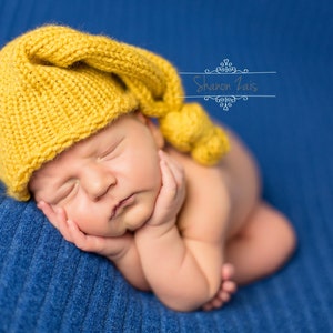 May include: A newborn baby wearing a yellow knitted cap with a pom-pom, resting on a blue textured blanket. The baby is sleeping peacefully, with hands gently supporting the head.