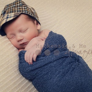 May include: A newborn baby boy is sleeping soundly in a blue knitted wrap. He is wearing a plaid patterned cap. The baby is lying on a beige textured surface.