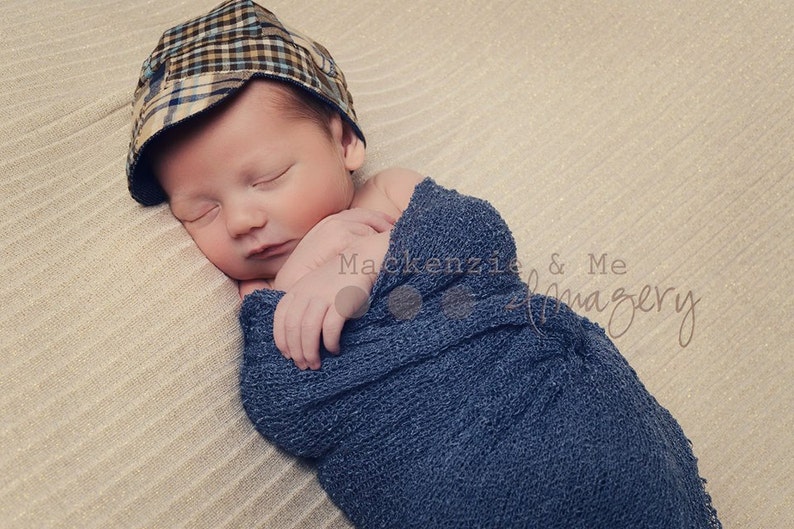 May include: A newborn baby boy is sleeping soundly in a blue knitted wrap. He is wearing a plaid patterned cap. The baby is lying on a beige textured surface.