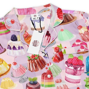 Puede incluir: Conjunto de pijama rosa claro con un patrón repetido de postres coloridos, que incluyen pasteles, gelatinas y pudines. El conjunto presenta una parte superior con cuello y una etiqueta que dice "Cabana Chocolate".