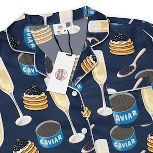 Könnte beinhalten: Marineblaues Pyjama-Set mit einem sich wiederholenden Muster aus Champagnergläsern, Kaviar-Dosen und Pfannkuchen mit Kaviar. Das Set enthält ein Oberteil mit Kragen und Tasche sowie eine weiße Paspelierung und ein Etikett mit der Aufschrift "Cabana Creations".