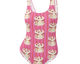 May include: One-piece swimsuit with a pink background and vertical red stripes. The swimsuit features a repeating pattern of white cakes topped with red cherries and cream-colored frosting. The straps and edges are pink.