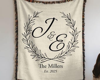 Puede incluir: Un tapiz tejido de color crema con un monograma negro de "J&E" rodeado de una corona de hojas. El texto "The Millers Est. 2025" está impreso debajo del monograma.