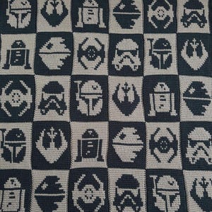 May include: A handmade, checkered blanket featuring Star Wars characters and symbols. The design includes images of R2-D2, a Stormtrooper helmet, and the Rebel Alliance symbol. The blanket is in shades of gray and black.