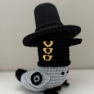 May include: A handmade crochet figure of a magpie wearing a black hat and coat with gold-colored buttons. The figure is primarily gray and black, with the text "KPOP Demon Hunter" and "Six-Eyed Magpie" in the image.