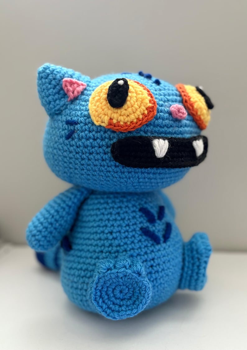 May include: A handmade blue crochet monster doll with orange, yellow, black, and pink accents. The doll has large eyes, a black mouth with white teeth, and blue stitched details. It is a whimsical, soft toy.