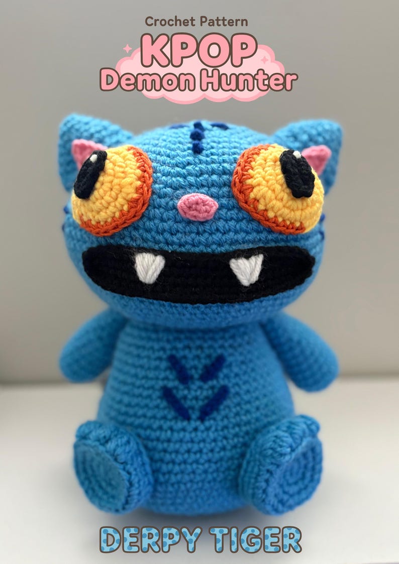 May include: A handmade blue crochet tiger with orange and yellow eyes, a black mouth, and white teeth. The text "KPOP Demon Hunter" and "Derpy Tiger" are displayed above and below the tiger. The crochet animal has small arms and legs.