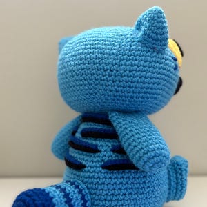 May include: A handmade blue crochet cat doll with black and blue stripes on its back and tail. The cat has small ears, a yellow eye, and a blue tail. The doll is made of soft yarn and is a cute, cuddly toy.