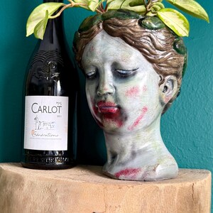 May include: A ceramic head planter with a zombie-like appearance, featuring green and gray tones with red accents on the lips and cheeks. The planter holds a green plant with light green leaves. A dark wine bottle is next to the planter.