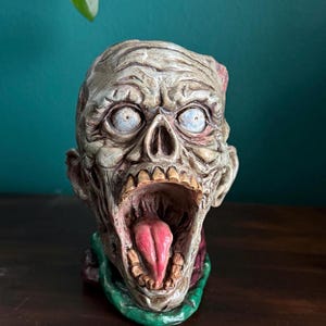 May include: A detailed zombie head sculpture with a gaping mouth and exposed tongue. The sculpture features a weathered, gray complexion with blue eyes and a green collar. The overall theme is horror and Halloween.