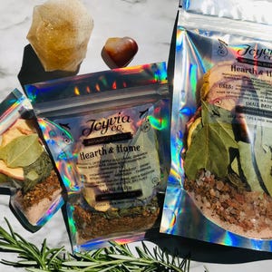 May include: Three holographic pouches filled with dried herbs and spices, labeled "Hearth & Home" by Joyvia Co. A quartz crystal and a heart-shaped stone are also visible. Rosemary sprigs are in the foreground.