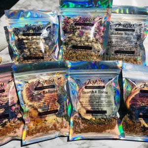 May include: Eight small, clear, iridescent bags filled with various dried herbs, flowers, and fruit slices. Each bag has a label with the name of a different blend, such as "Goddess Glow", "Solar Radiance", and "Sacred Heart".