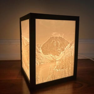 May include: A black framed lamp with a white, illuminated image of a mountain range and a lake. The image is 3D and appears to be carved into the surface of the lamp.