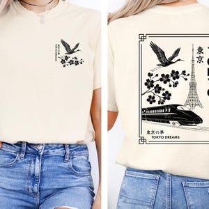 May include: A cream colored t-shirt with a black graphic design featuring a crane, cherry blossoms, a train, and the Tokyo Tower. The text on the shirt reads "Tokyo Dreams" and "東京の夢".
