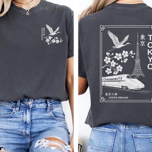 May include: A dark gray t-shirt with a white graphic print of a crane, cherry blossoms, a train, and the Tokyo Tower. The text "TOKYO DREAMS" is printed below the graphic.
