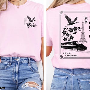 May include: Pink t-shirt with a black graphic design featuring a crane, cherry blossoms, a Tokyo Tower, and a bullet train. The text "TOKYO" and "東京" are also included in the design. The text "東京の夢 TOKYO DREAMS" is at the bottom of the design.