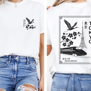 May include: White t-shirt with a black and white graphic print of a crane, cherry blossoms, and a train in front of the Tokyo Tower. The text "TOKYO" is written vertically on the right side of the graphic. The text "東京の夢 TOKYO DREAMS" is written below the graphic.