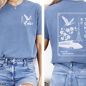 May include: A blue t-shirt with a white graphic design featuring a crane, cherry blossoms, a train, and the Tokyo Tower. The text on the shirt reads "TOKYO DREAMS" and "東京".