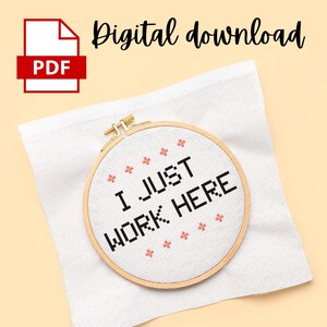 Puede incluir: Un aro de bordado blanco con un diseño de punto de cruz negro que dice "I JUST WORK HERE" rodeado de flores rosas. El aro está sobre un fondo de tela blanca.