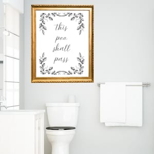May include: A framed wall art print with the text "this poo shall pass" in a bathroom setting. The gold-colored frame surrounds a white background with black botanical illustrations.