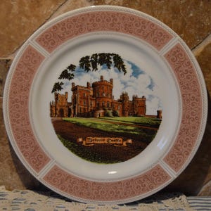 May include: A decorative plate featuring a detailed illustration of Belvoir Castle. The plate has a white center with a scenic image of the castle, framed by a pink border with an ornate design. The text "Belvoir Castle" is below the castle image.