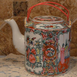 Puede incluir: Una tetera de porcelana blanca con asa y tapa rojas. La tetera está decorada con coloridos diseños florales y figurativos en rosa, verde y amarillo. El pico y los accesorios del asa tienen detalles dorados. La tetera es probablemente vintage.