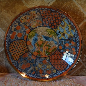 May include: Round decorative tray with a gold rim and a patterned design. The tray features a central image of two blue birds on a branch, surrounded by various colorful patterns in orange, blue, and yellow.