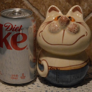 May include: A ceramic cat figurine with a cream-colored head, orange-spotted nose, and blue painted pants, positioned next to a 12 fluid ounce Diet Coke aluminum can for scale. The cat has a wide, smiling expression and a small tail visible on the side.