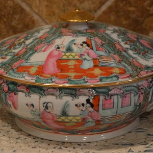 May include: A white porcelain serving dish with a lid, featuring a colorful hand-painted design. The design includes figures in pink and blue robes, set against a backdrop of trees and buildings. The dish has gold trim and handles.