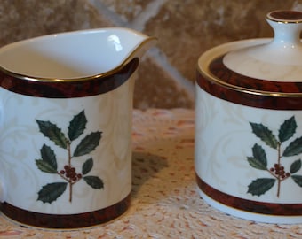 May include: Creamer and sugar bowl set with a festive holly design. The white ceramic pieces feature a burgundy band and gold trim. A holly branch with green leaves and red berries decorates each piece, perfect for holiday gatherings.