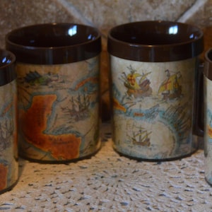 May include: Four brown-handled coffee mugs with a map design. The mugs have a brown rim and handle, and the body of the mugs feature a vintage map design with ships and nautical elements. The mugs are arranged on a white lace doily.