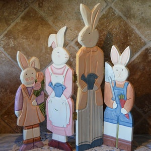 Op de afbeelding: Vier houten paashaasfiguren. Elke konijn is geschilderd met pastelkleuren en gekleed in verschillende outfits, waaronder overalls, schorten en jassen. Eén konijn houdt een boeket bloemen vast, een ander een gieter en een derde een schop en een wortel.