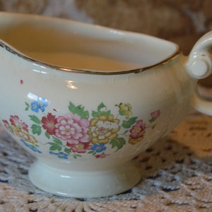 Peut inclure: Saucière crème avec un motif floral composé de fleurs roses, rouges, bleues et jaunes. Le bateau a une bordure dorée, une anse et un socle. Une pièce classique pour servir les sauces et les jus.
