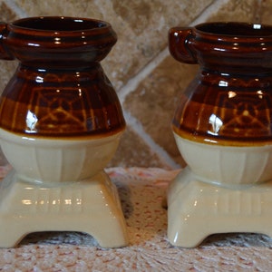 May include: Two brown and white ceramic mugs shaped like wood-burning stoves. The mugs have a handle on the side and a base with three legs.