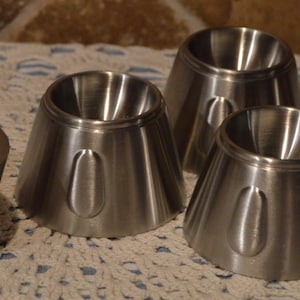 Puede incluir: Un conjunto de cinco hueveras de metal cepillado y una tapa a juego. Las hueveras tienen una forma cónica ensanchada con un asa redondeada. La tapa tiene una forma cilíndrica sólida. Los artículos están dispuestos sobre un mantel de encaje blanco.