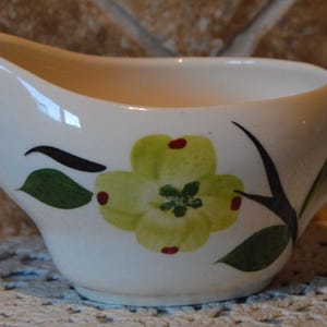 May include: A cream-colored ceramic gravy boat featuring a painted design of a light green dogwood flower with dark green leaves and small red accents. The vessel has a curved pouring spout on one side and a sturdy handle on the opposite side.