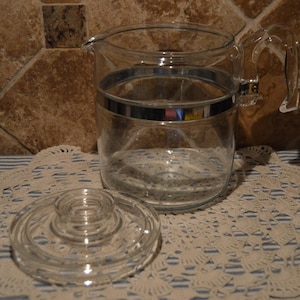 以下が含まれることがあります： 中央にシルバーのバンドと、お揃いの蓋が付いた透明なガラスピッチャー。ピッチャーにはハンドルが付いており、白いレースのドイリーの上に置かれています。背景は茶色のタイルです。