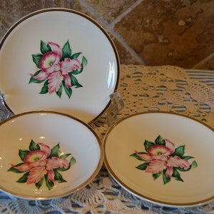 May include: Three white ceramic plates with gold trim and a floral design. The plates feature pink and green orchids.