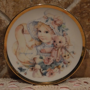 Puede incluir: Un plato de porcelana con un borde dorado presenta una pintura de una niña con un vestido y un sombrero blancos que sostiene un oso de peluche. La niña está rodeada de flores rosas y blancas. El plato está firmado "Jan Hagara 1996".