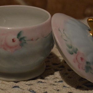 May include: A small, open, porcelain trinket dish with a matching lid. The dish and lid are painted with pink roses and green leaves on a pale blue background. The lid has a gold-colored handle.