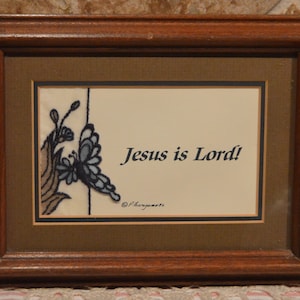 May include: A framed artwork with a brown wooden frame and a beige mat. The artwork features a blue butterfly with a floral design and the text "Jesus is Lord!" in black lettering.