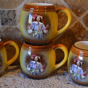 Może przedstawiać: Zestaw czterech żółtych i brązowych ceramicznych kubków z wzorem mężczyzny w niebieskim i fioletowym stroju. Każdy kubek ma inny wzór mężczyzny w innej pozie.