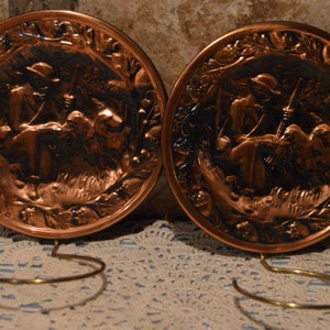 May include: Two copper wall plaques with a raised design of a person with two dogs. The plaques are hanging on a wall with brass hooks.