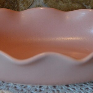 May include: A light pink, oval-shaped ceramic bowl with a scalloped rim. The bowl has a smooth, matte finish and sits on a decorative doily. The bowl is empty and appears to be vintage.